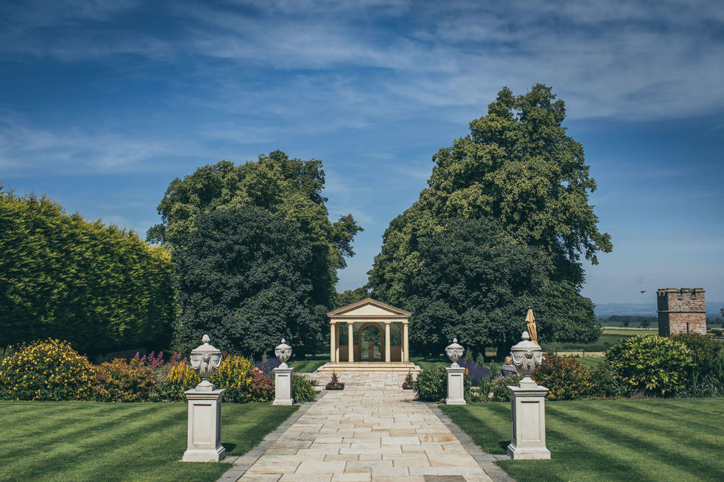 Rowton Castle's gardens are in full bloom. It is a blue sky day, the lawns have been striped, in the middle the patio and decorative pillars lead to the Linden Belvedere
