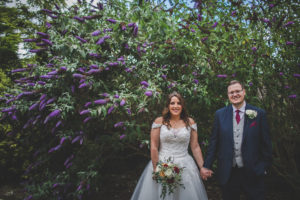 A Rowton Castle Bride and Groom photographed hand in hand in front of the purple blooming buddleja