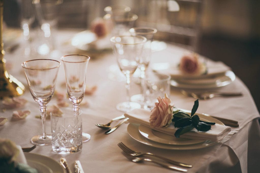 Close up of the wedding breakfast table with white linen, gold cutlery and gold rimmed wine glasses. A pink rose sits at each place setting