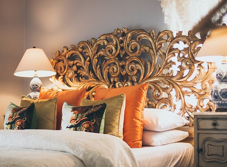 The bed in the Lady Charlotte Room (Tower House) is photographed up close. It's intricate gold headboard and orange and gold cushions are the focus