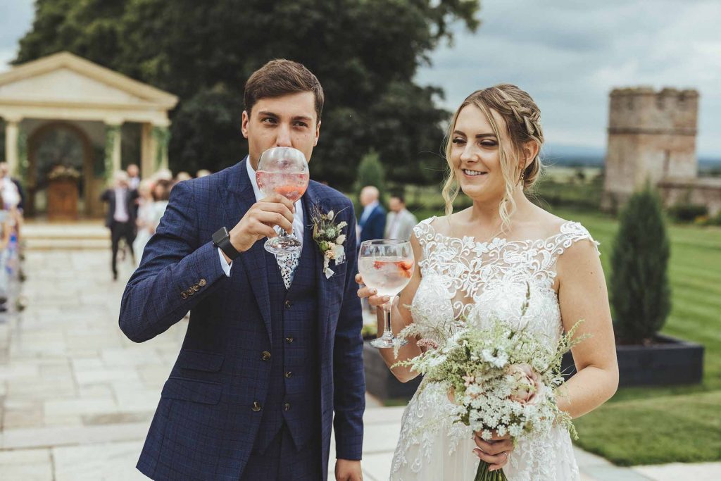 Bride and groom take the first sip of their reception drink following their outdoor wedding ceremony. Guests are seated in rows behind them
