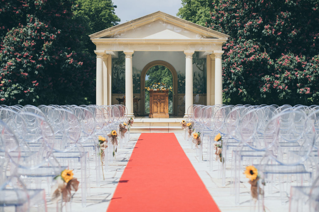 The linden belvedere is dressed for an outdoor ceremony; red aisle carpet and acrylic chairs are placed in rows