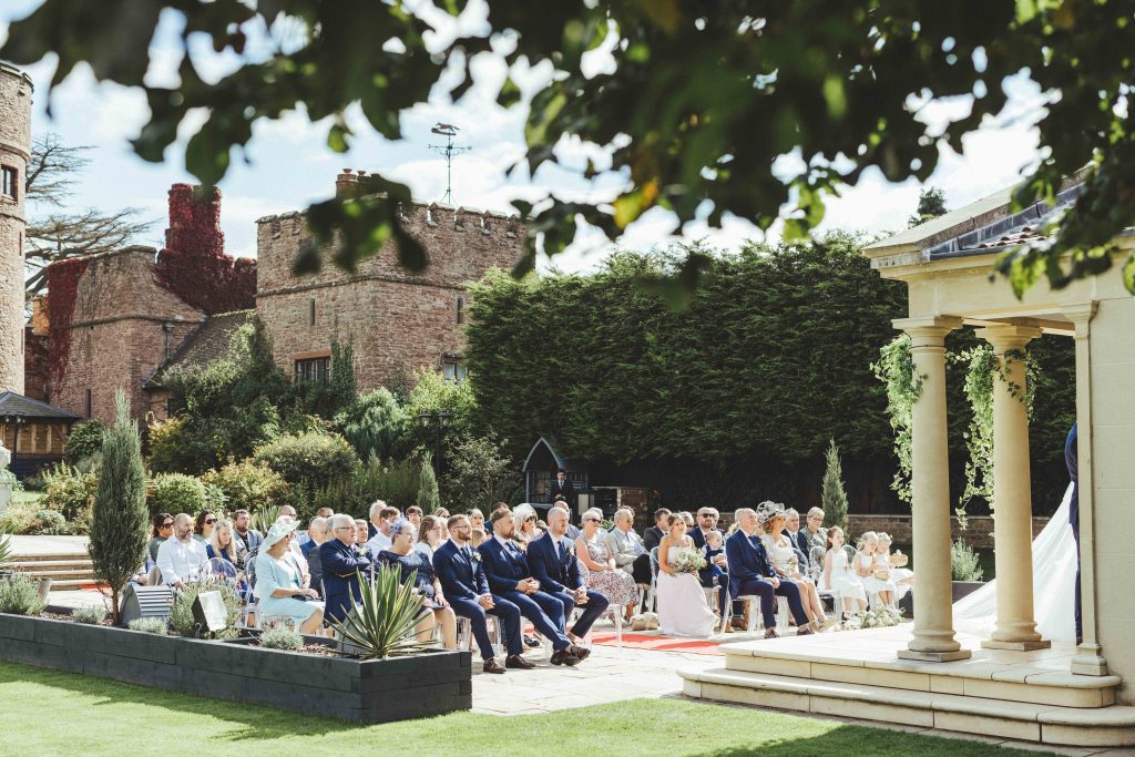 The outdoor ceremony is just beginning. Guests are sat in rows and the castle turrets can be seen above