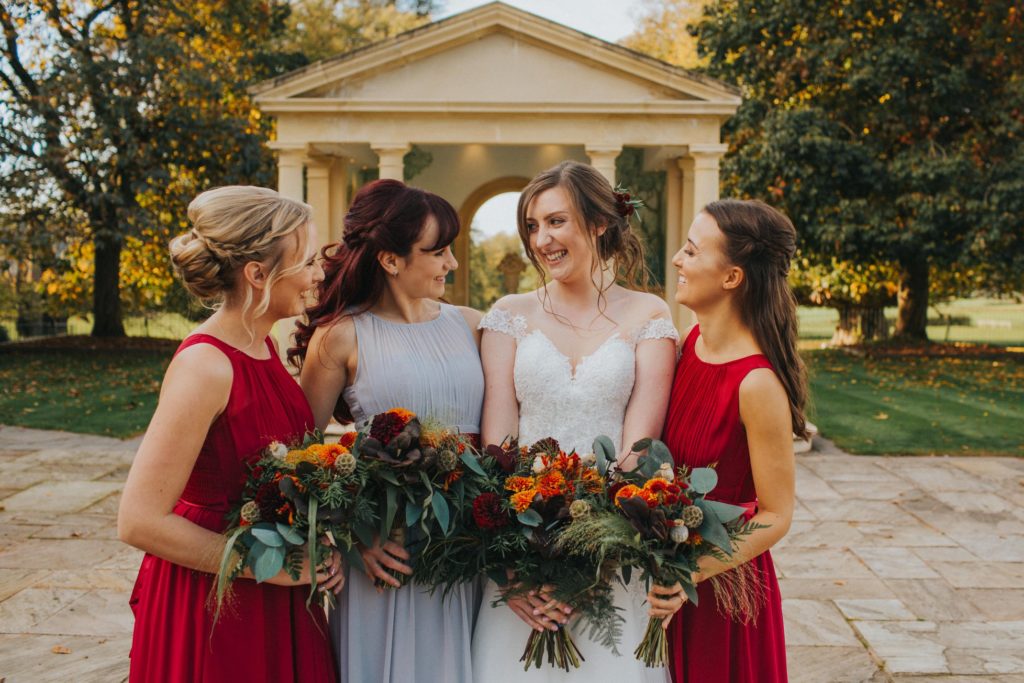 The bride and her bridesmaids are photographed laughing in front of the linden belvedere. The bridesmaids wear red and hold bouquets of autumnal flowers