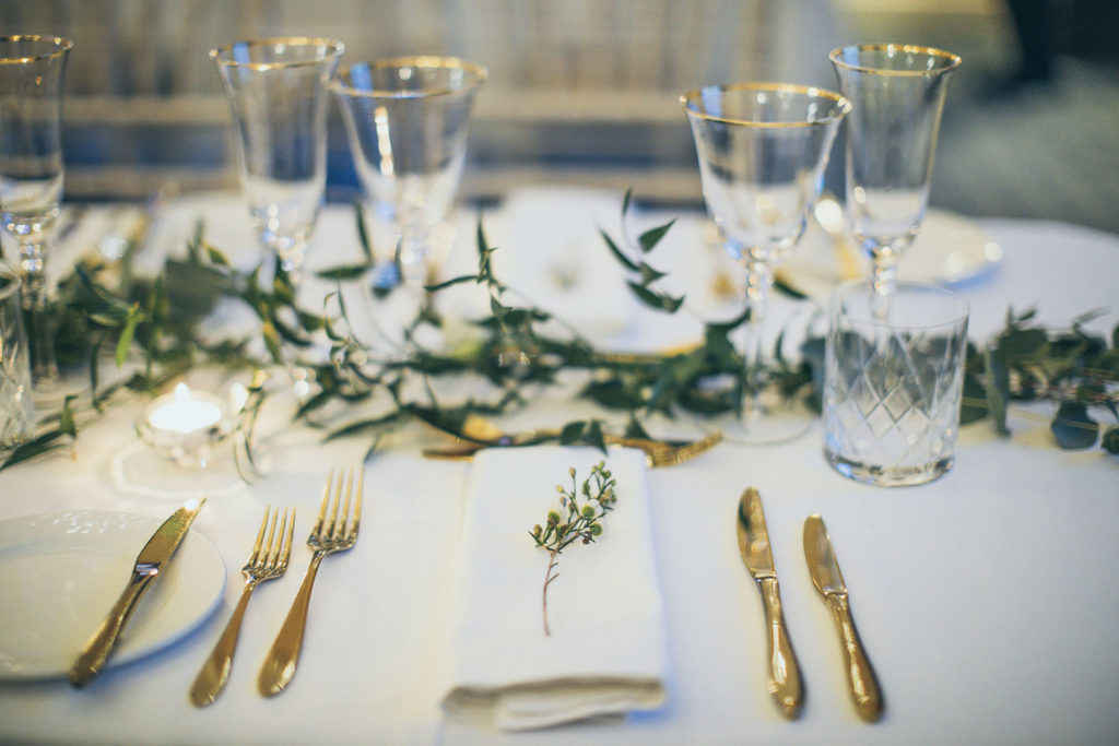 A close up of a place setting. White napkin, gold cutlery and gold rimmed glasses. A single piece of wax flower sits upon the napkin