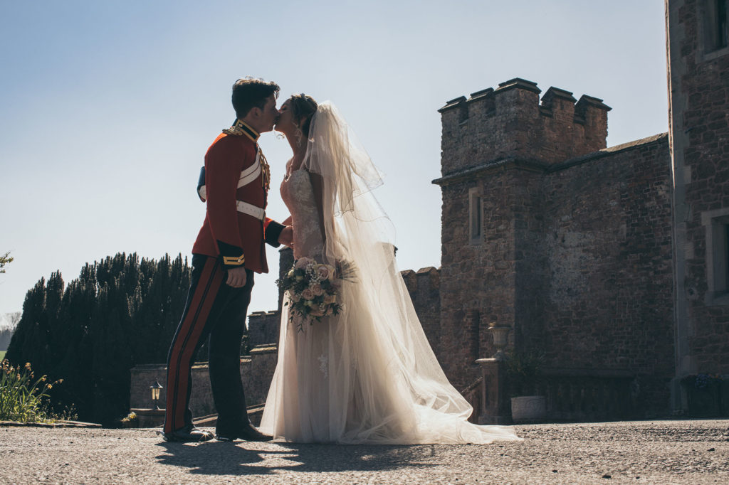 The bride and groom stop and kiss at the front of the castle. The groom wears his military uniform and the bride a lace gown and full veil