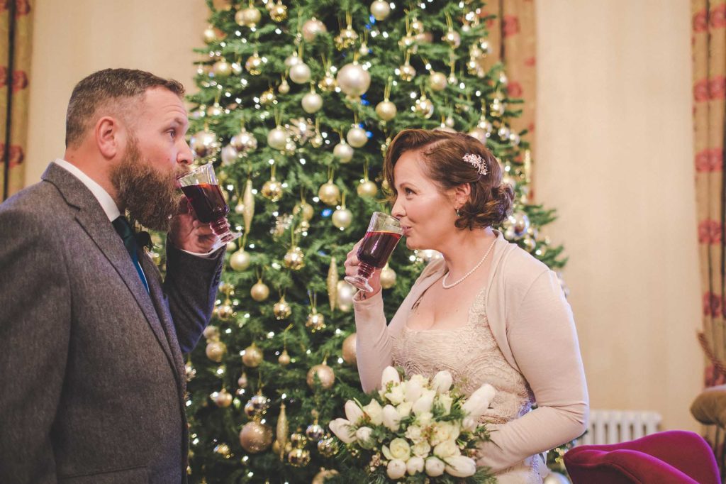 The bride and groom sip a reception drink of mulled wine in front of the lounge christmas tree