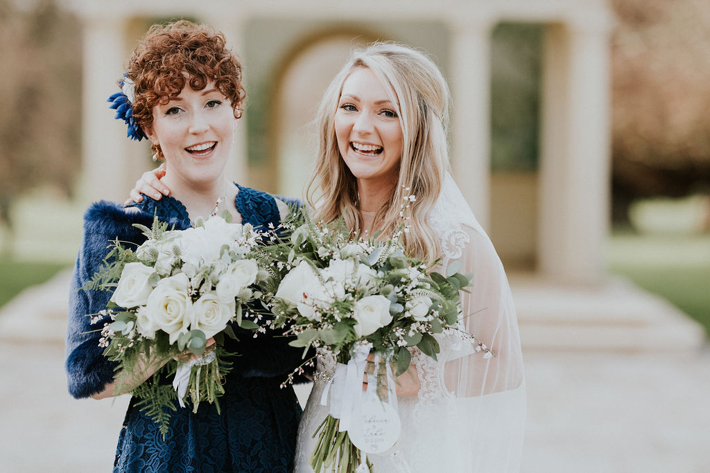 A bride (dressed in lace) and her maid of honor (dressed in navy blue) are captured on a winters day in front of the linden belvedere. Both are laughing and holding bouquets