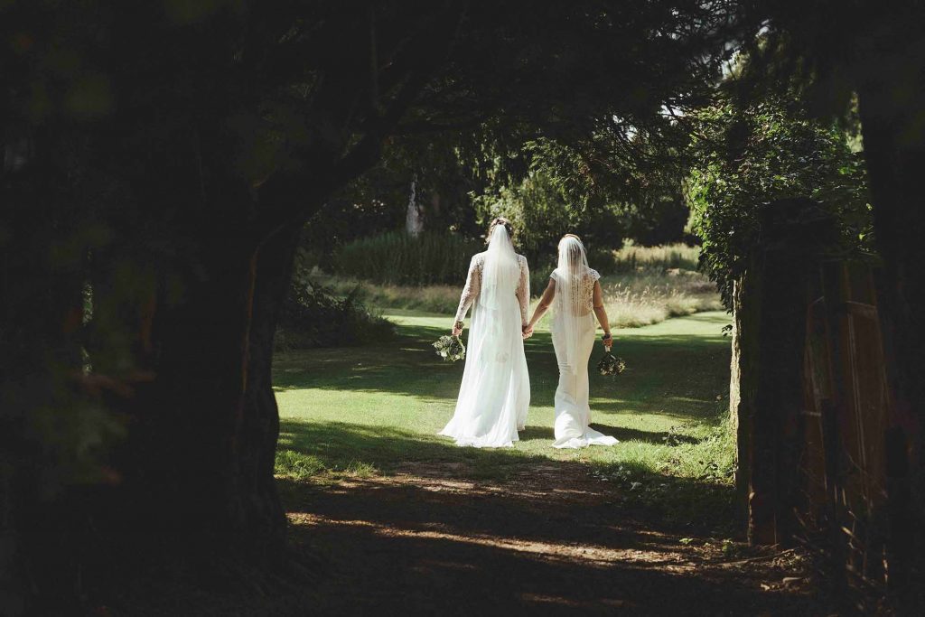 The brides walk hand in hand (both holding their bouquets) as they wonder through the trees further in the grounds