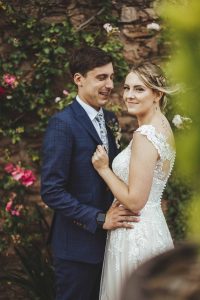The lovely Samantha and Lewis (bride and groom) are photographed up close in front of the rose wall. Pink roses are in bloom