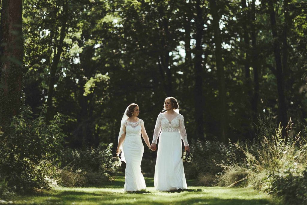 The brides walk hand in hand through the trees in the grounds of Rowton Castle