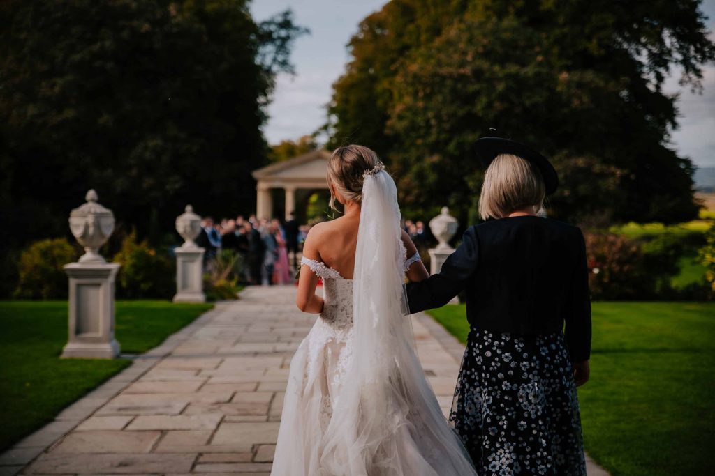 The bride is given away by her mother as guests look back in this outdoor wedding ceremony