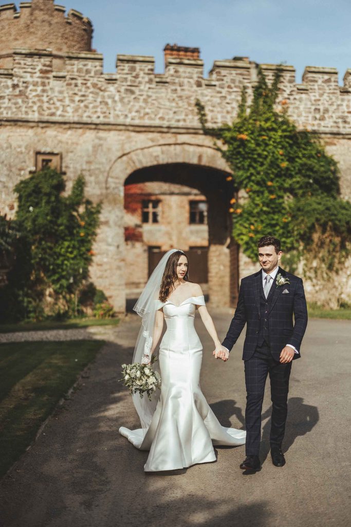 Emillie & Richard (Bride and Groom) walk hand in hand toward the castle woodland, the turreted archway can be seen in the background
