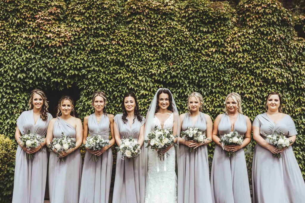 Bride Esther and her 7 bridesmaids are photographed outside of Tower House in front a wall of green virginia creeper. Each holds a bouquet, the bridesmaids wear silver