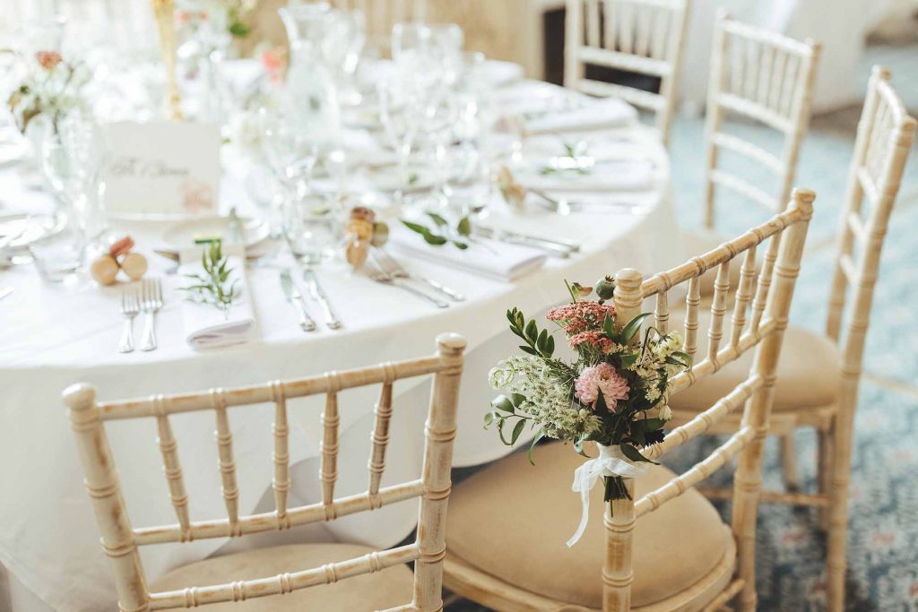 A close up of the wedding breakfast table, limewash chiavari chairs dressed with florals and pastel macaroon favours feature