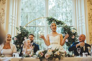 The bride stands in front of a floral installation at her top table to make a speech of her own. Her groom, mother, father and daughter all have smiles on their faces