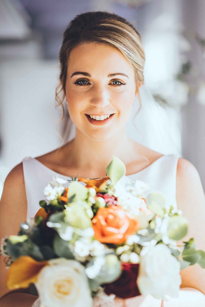 Smiling bride Hollie is photographed in the Orchard Suite holding her bouquet that is filled with autumnal coloured roses