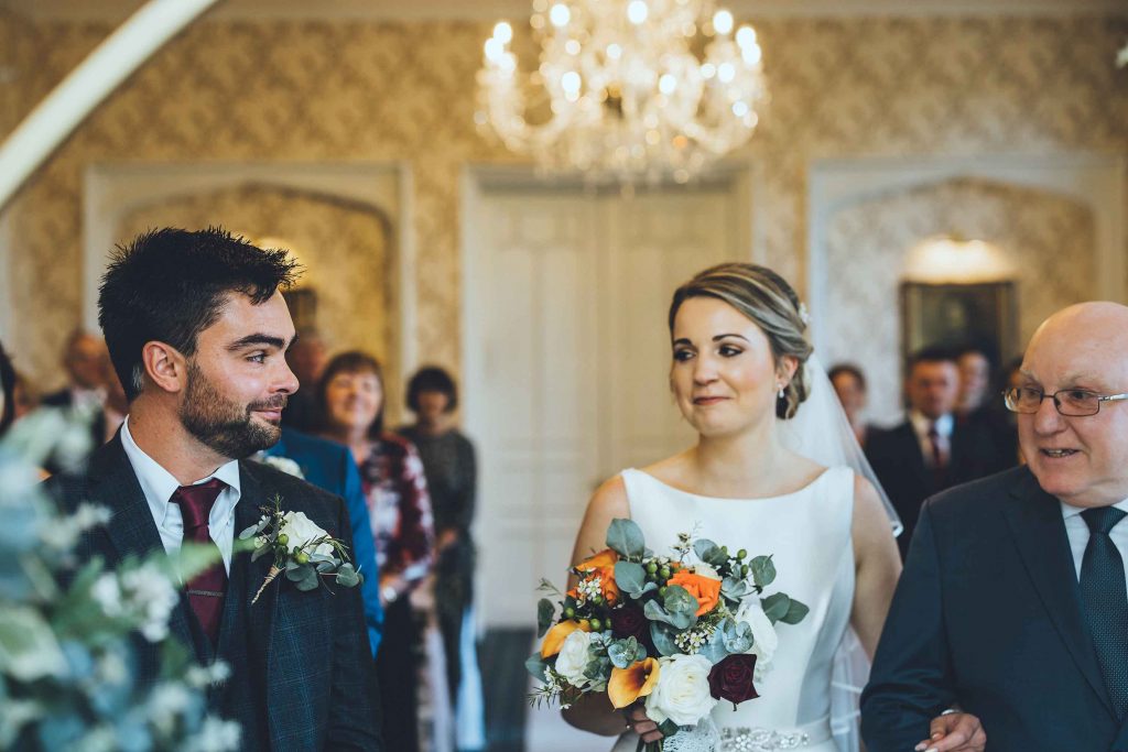 Bride Hollie is walked down the aisle by her proud father., she carries a bouqet that contains autumnal flowers. Groom Callum is noticeably emotional