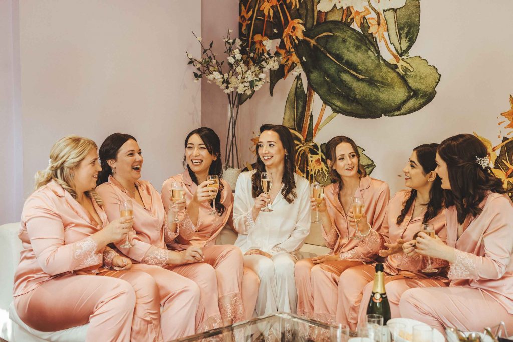 Bride Emilie and her six bridesmaids sit on the sofa in the orchard suite wearing silk pj sand drinking champagne