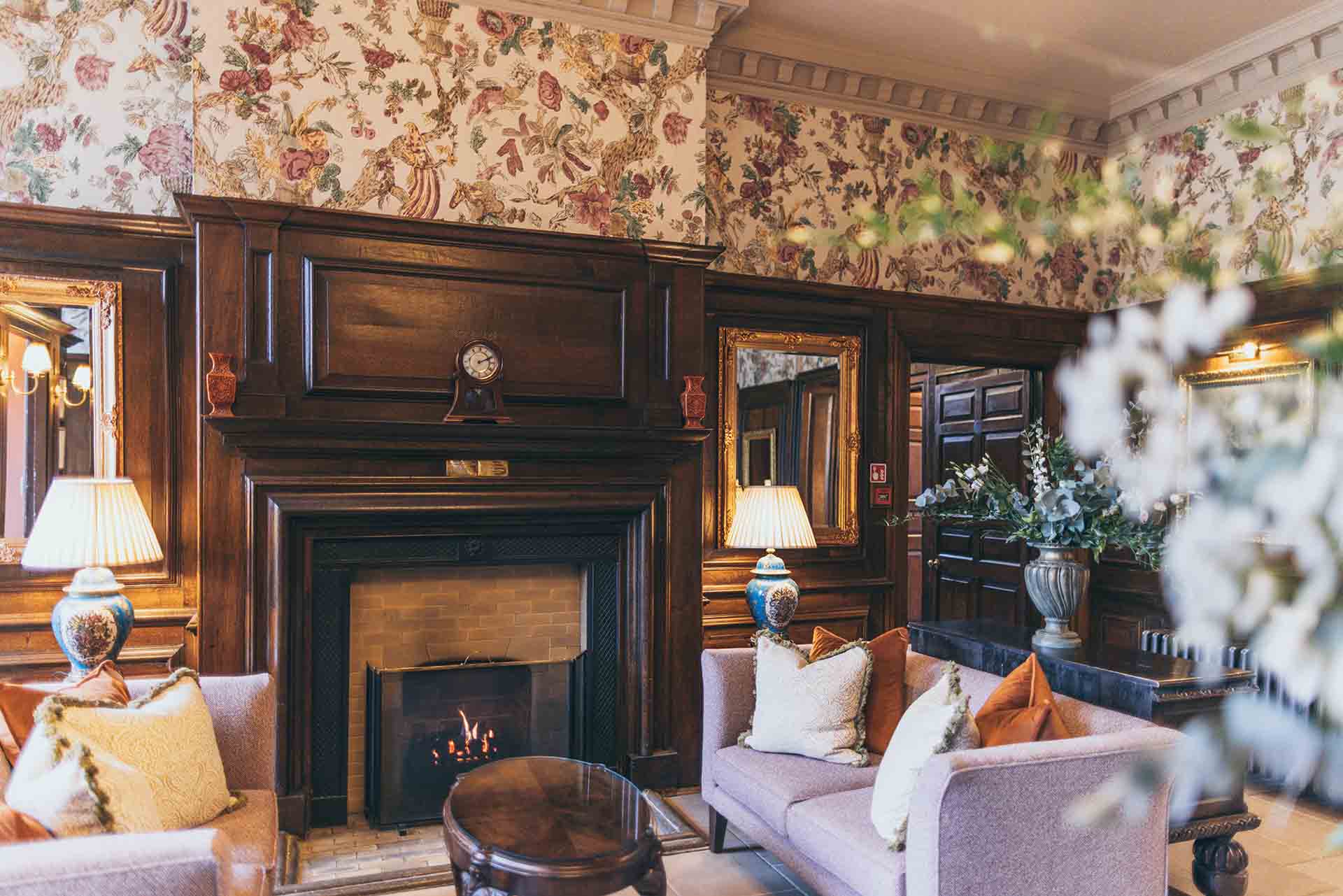 Cosy vintage-style reception room with a wooden fireplace mantel, floral wallpaper, upholstered armchairs with cushions, table lamps, mirrors, and decorative flowers.