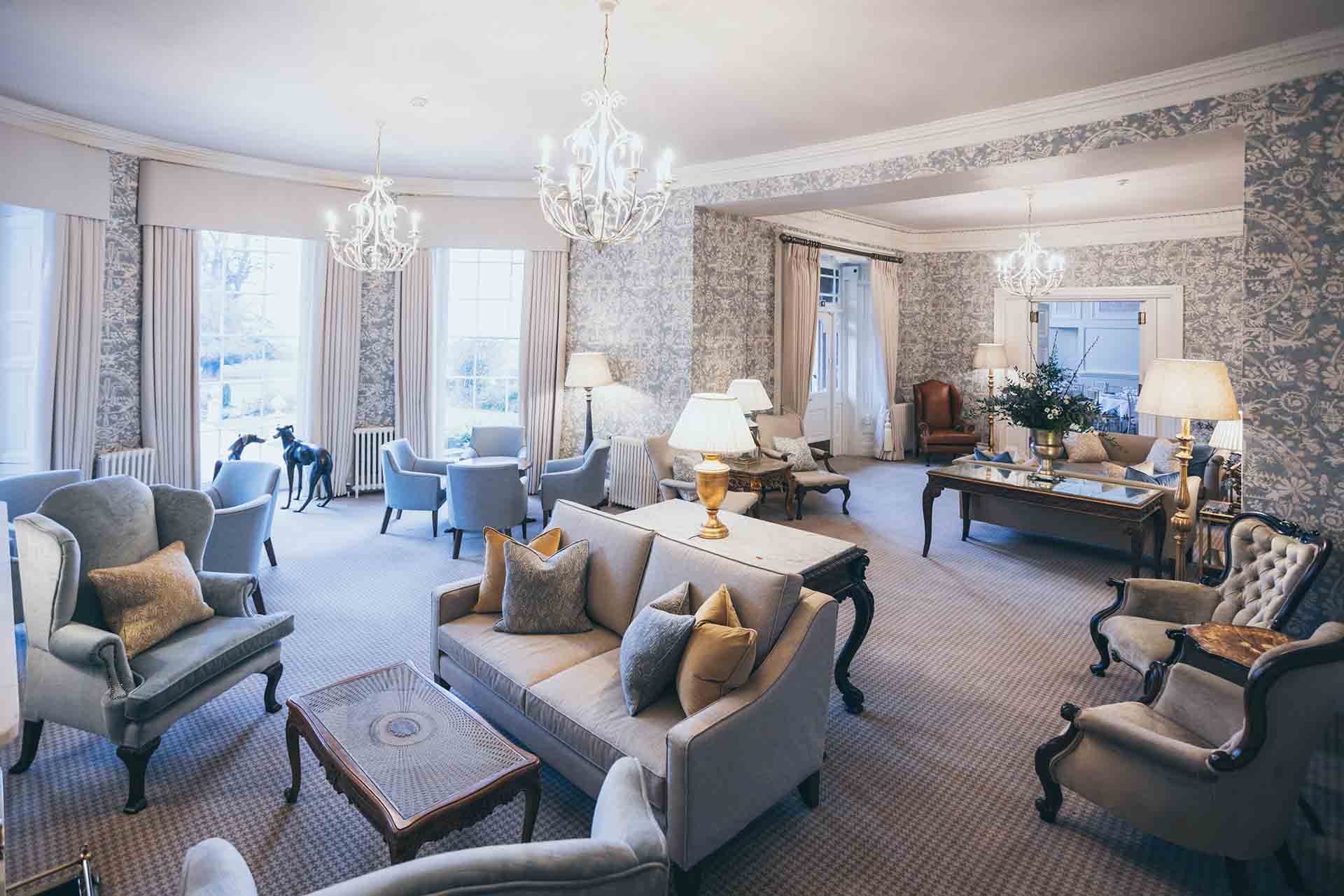 Spacious elegant lounge with chandeliers, upholstered seating, side tables with lamps, and large windows overlooking the gardens in a historic castle interior.