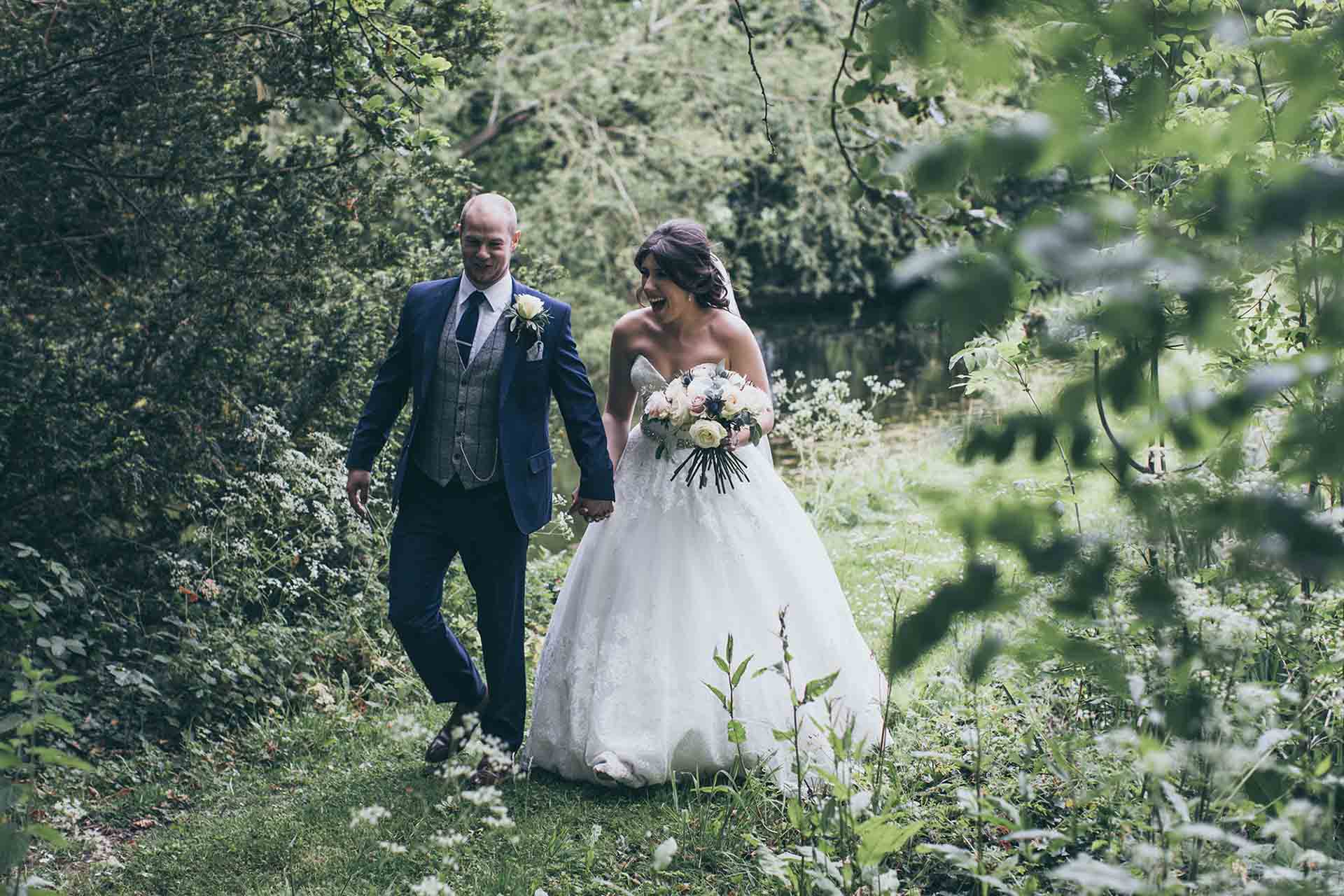 A newlywed couple holding hands and walking through a leafy woodland path at Rowton Castle, surrounded by lush greenery on their wedding day.