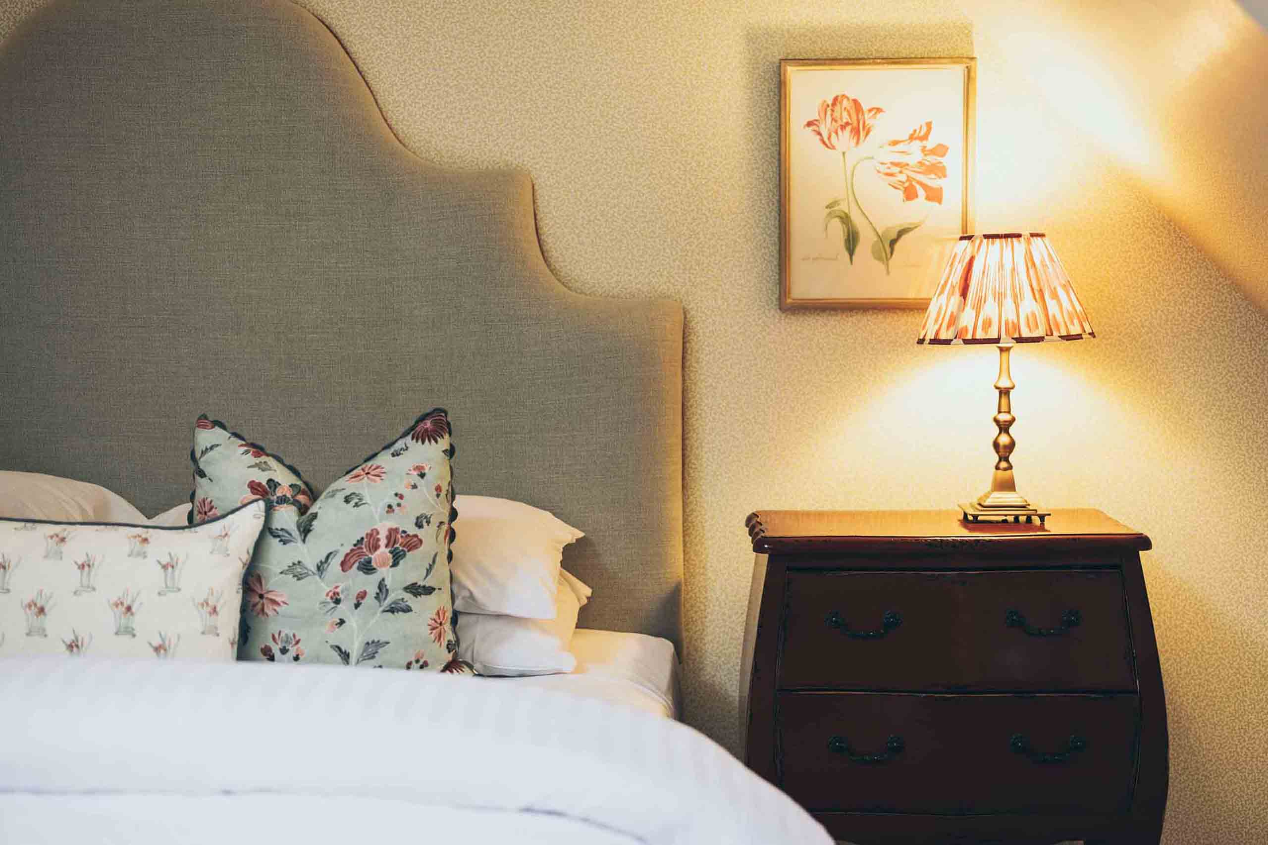 Castle bedroom detail featuring a softly upholstered headboard, floral cushions, and a bedside table illuminated by warm lamplight.
