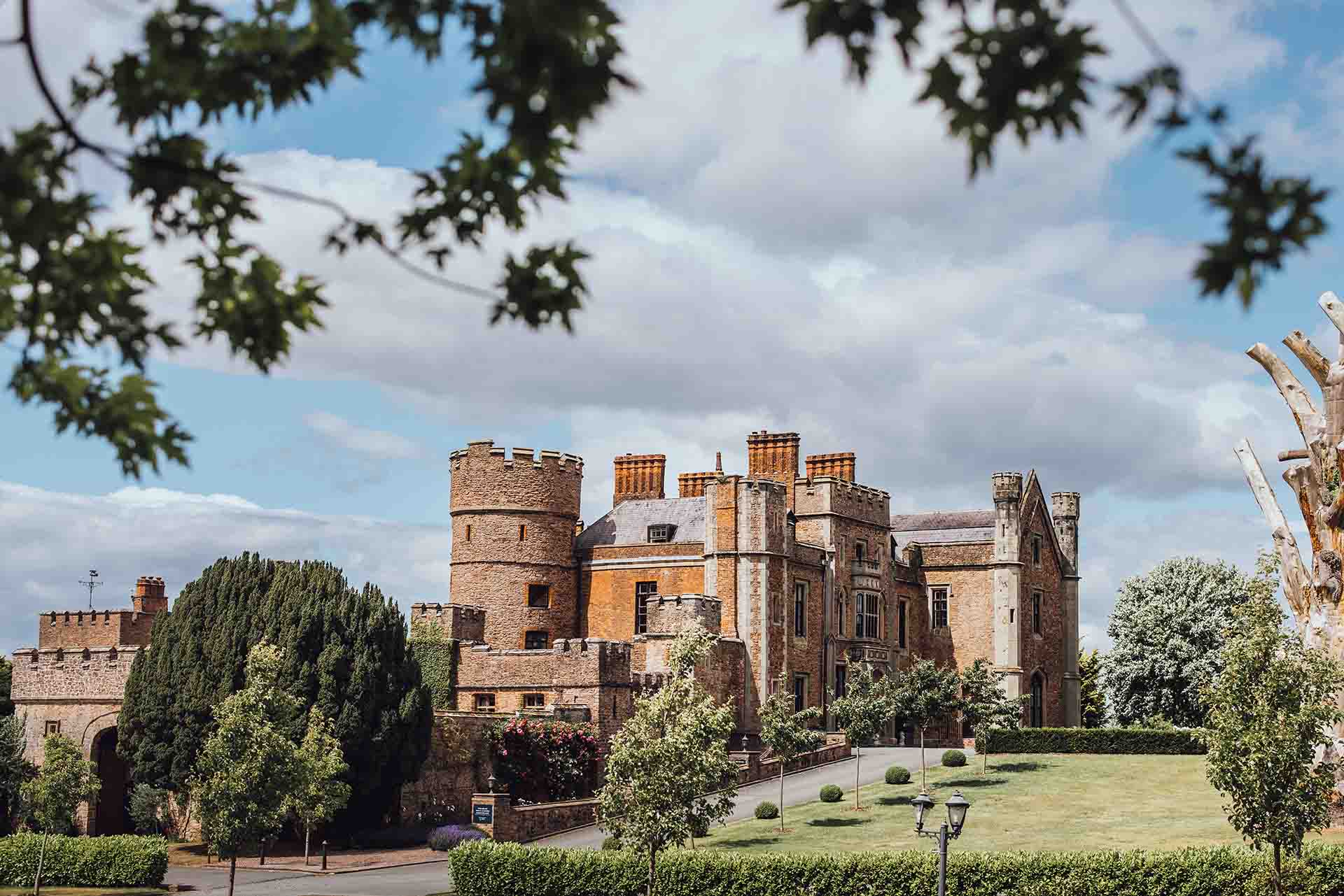 A historic sandstone castle with round towers and tall chimneys, set on a manicured lawn with trees and hedges, under a partly cloudy blue sky.