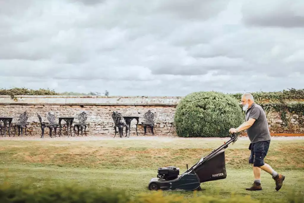 An older man with a gray beard pushes a lawn mower across a neatly trimmed grassy lawn in a garden, with a stone wall, patio tables and chairs, and a large round shrub under a cloudy sky.