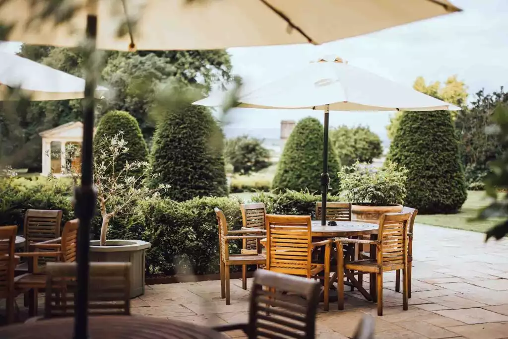 Outdoor garden terrace with wooden tables and chairs under large cream umbrellas, surrounded by manicured hedges and cone-shaped topiary trees in a landscaped countryside setting.