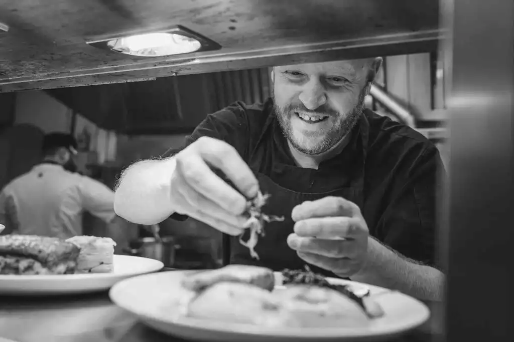 Chef carefully garnishing a plated dish in a professional kitchen, smiling as he adds finishing touches.