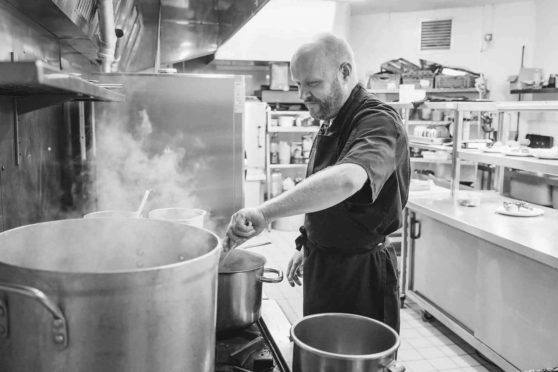 Chef stirring a pot on a commercial stove in a busy restaurant kitchen with steam rising.
