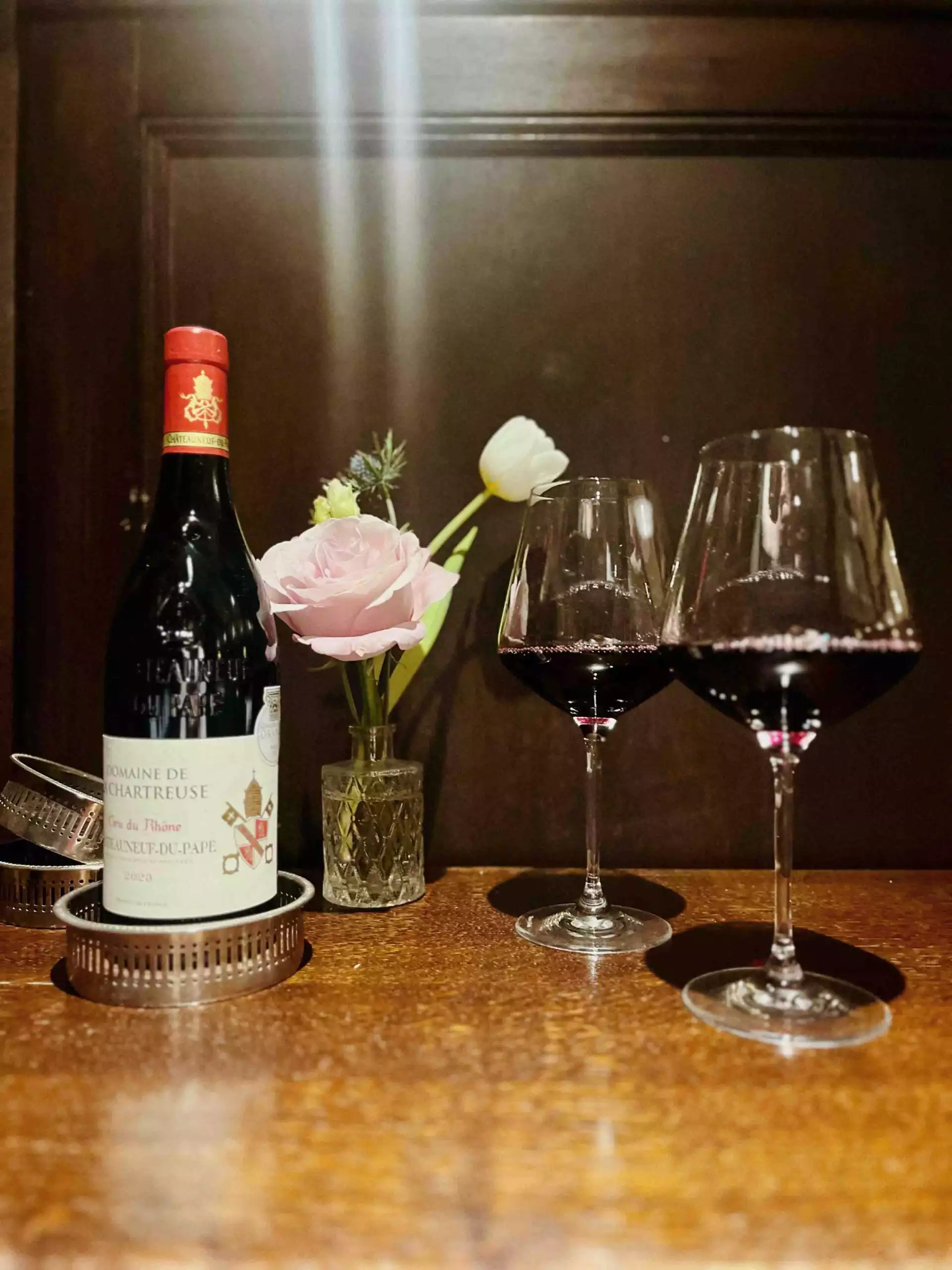A bottle of red wine beside a small vase of pale pink and white flowers, with two partially filled red wine glasses on a polished wooden table against a dark panelled background.