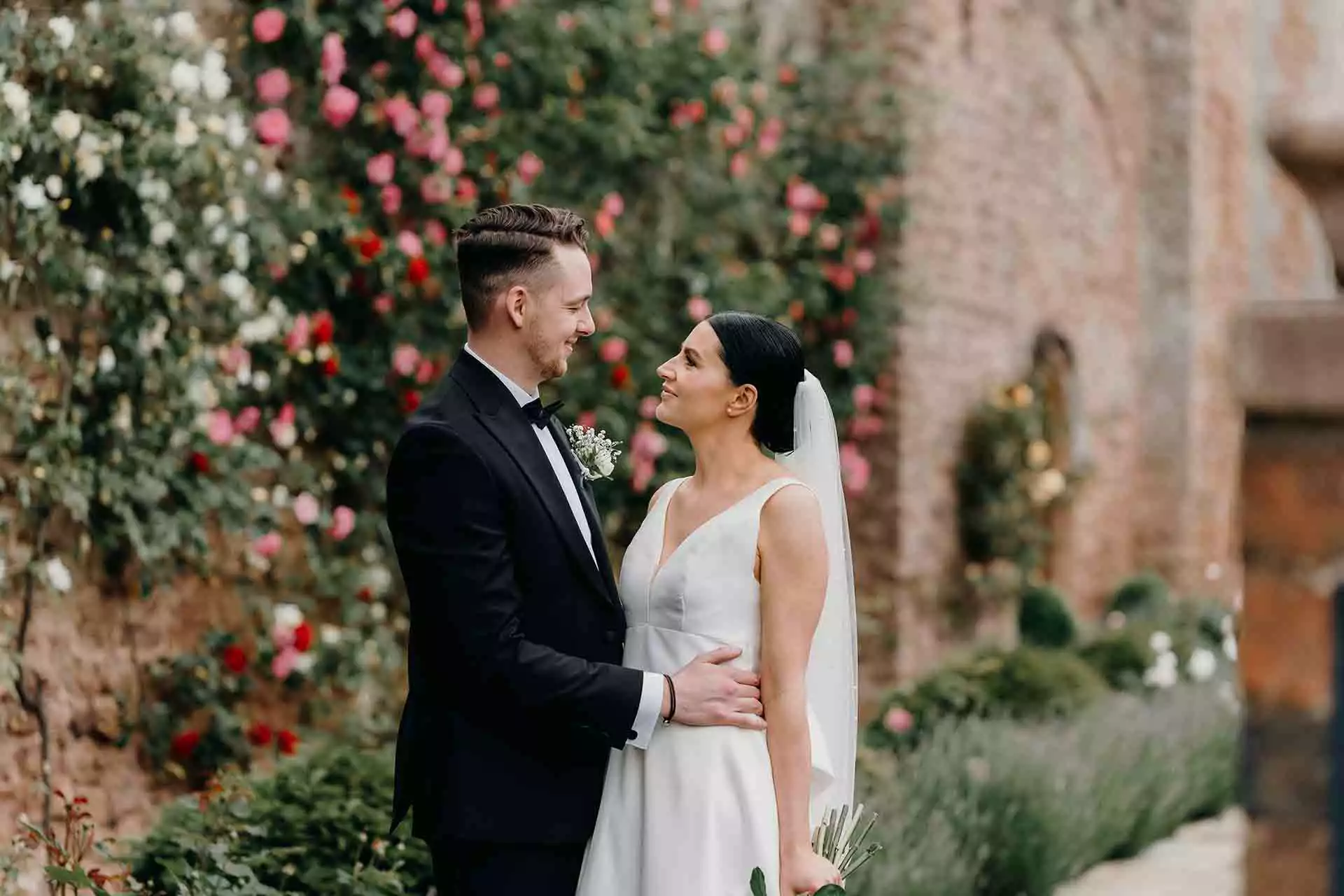 Bride and groom embracing in front of blooming flowers and historic brick walls at Rowton Castle.
