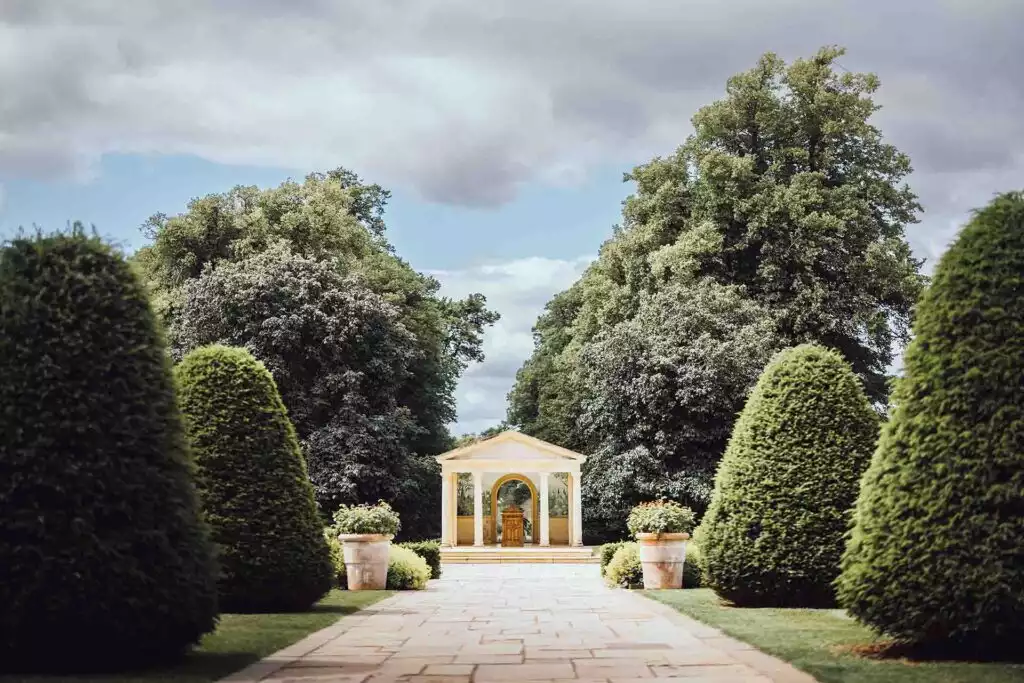 A tree-lined garden pathway leading to the classical outdoor ceremony pavilion at Rowton Castle, framed by manicured topiary and stone planters.