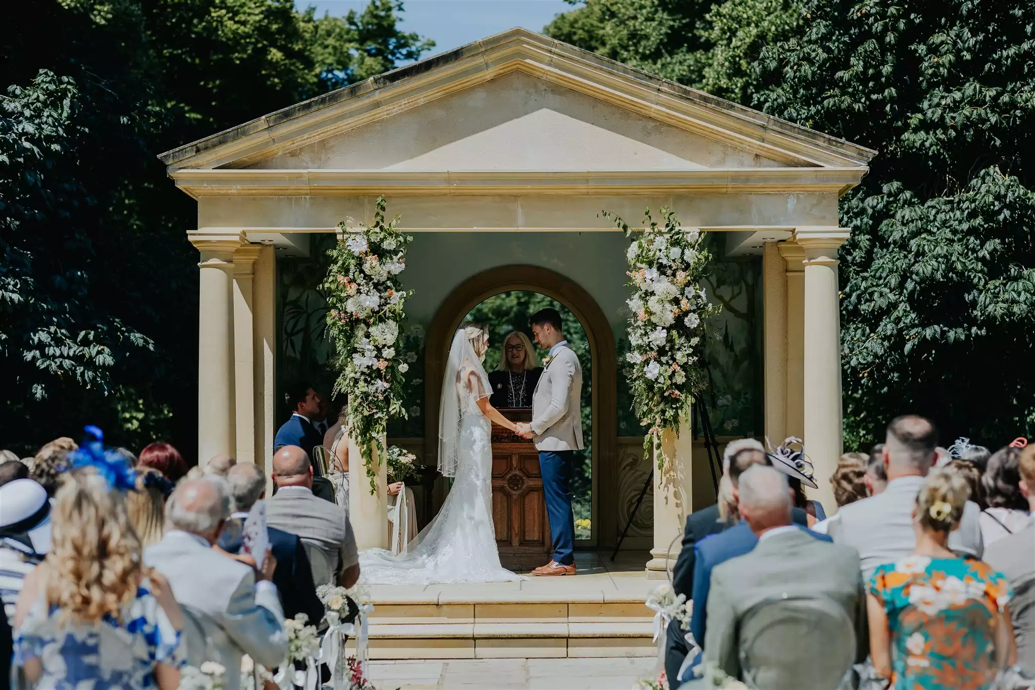 Bride and groom exchanging vows during an outdoor wedding ceremony beneath a classical stone pavilion adorned with floral arrangements, surrounded by seated guests in a garden setting.