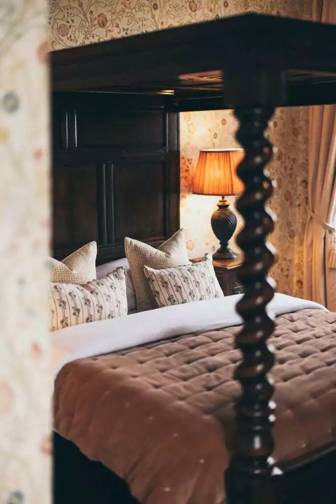 A cosy four-poster bed dressed in soft linens and patterned cushions, set within a warmly lit castle bedroom with floral wallpaper and classic furnishings.
