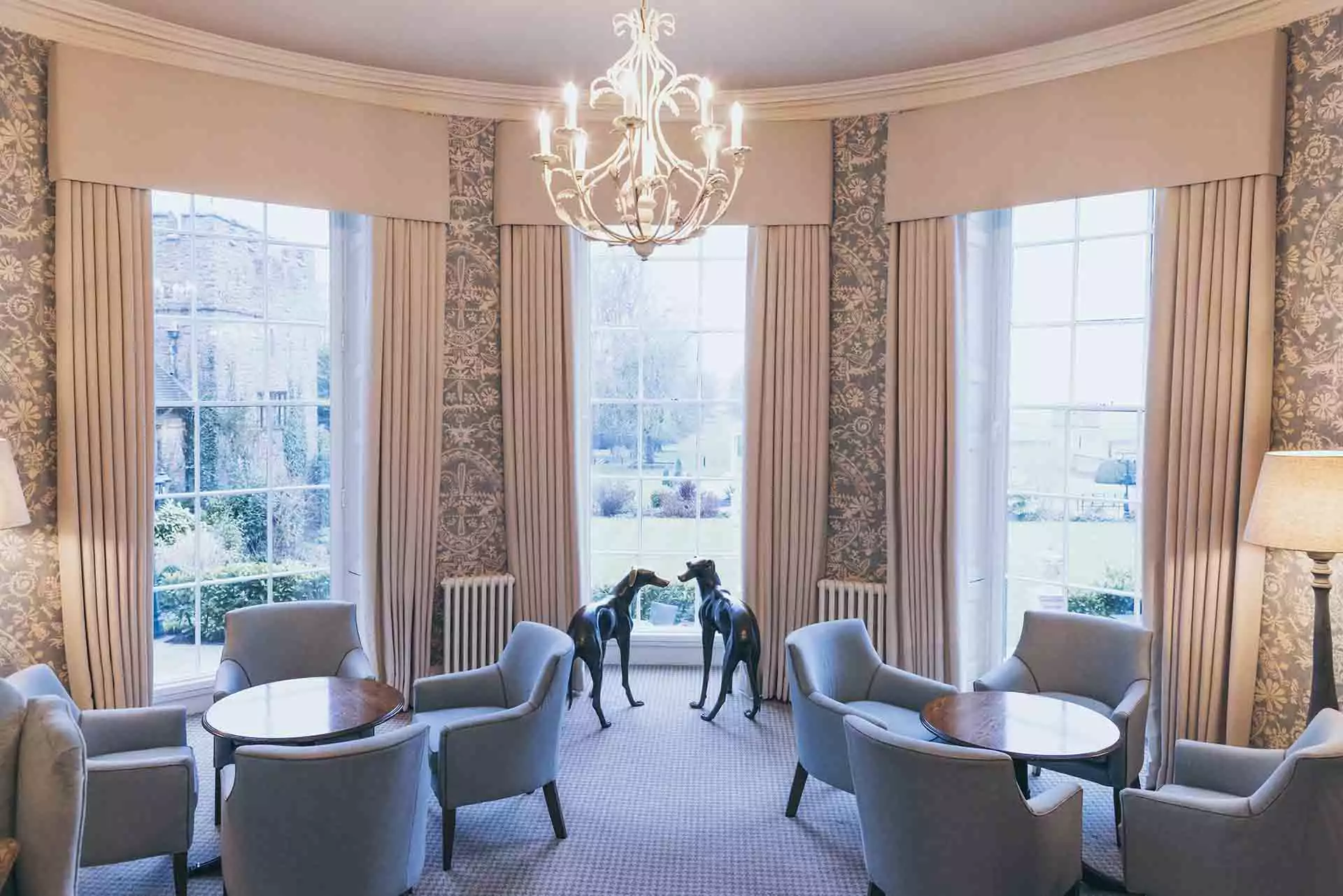 Bright and elegant lounge area at Rowton Castle featuring tall bay windows overlooking the gardens, a central chandelier, upholstered armchairs with round tables, and decorative greyhound statues.