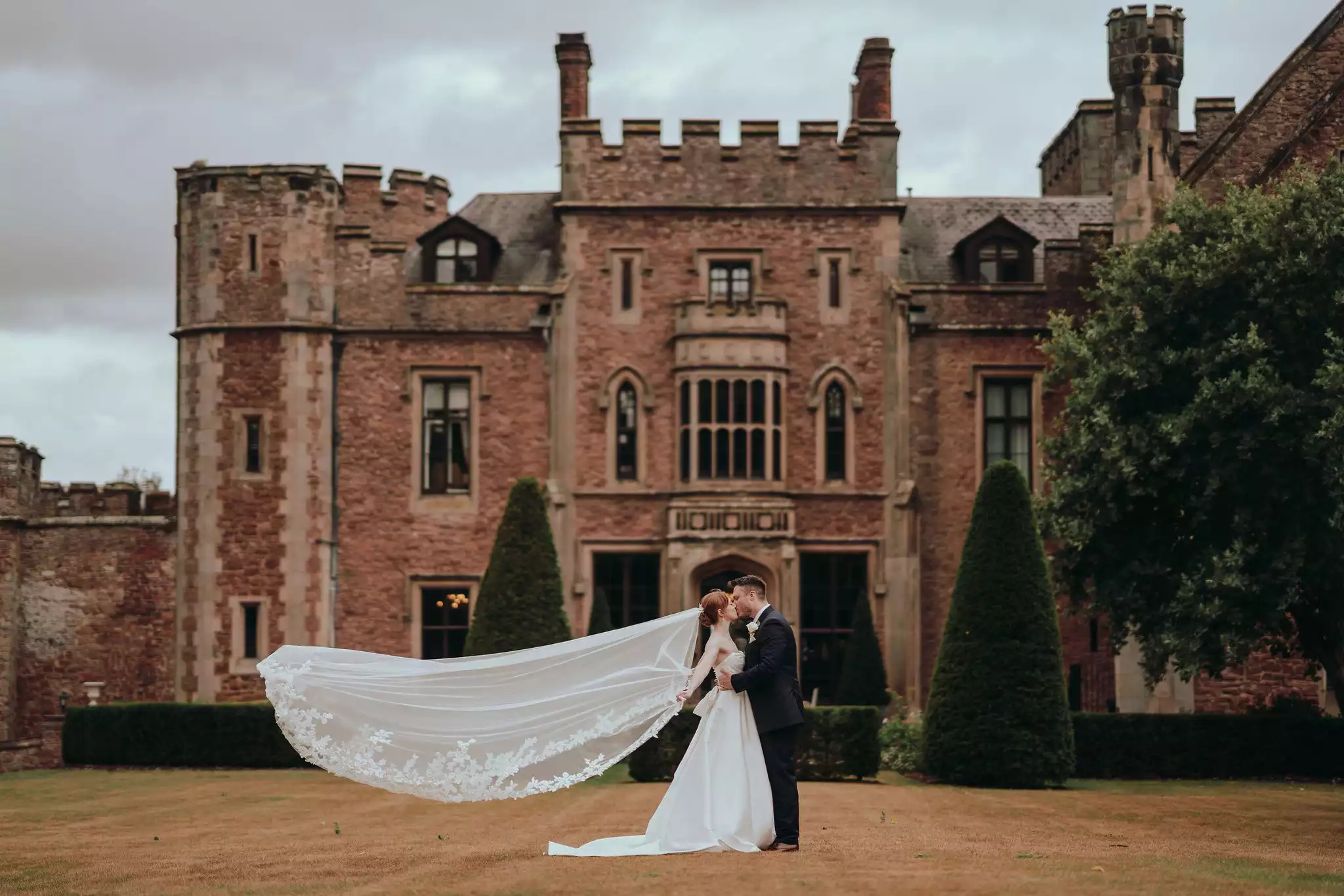 Bride and groom sharing a kiss in front of a historic castle wedding venue, with the bride’s long veil flowing across the lawn.