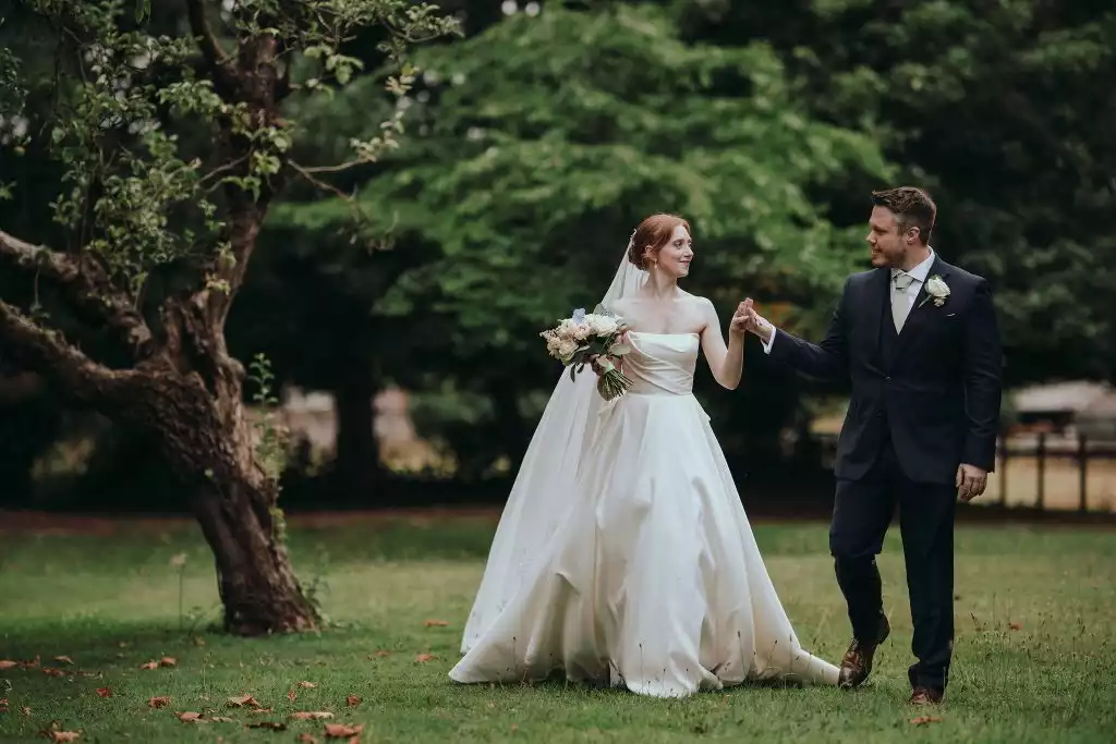 Bride holding a bouquet of blush roses walking hand in hand with the groom through lush green gardens at a countryside wedding venue.