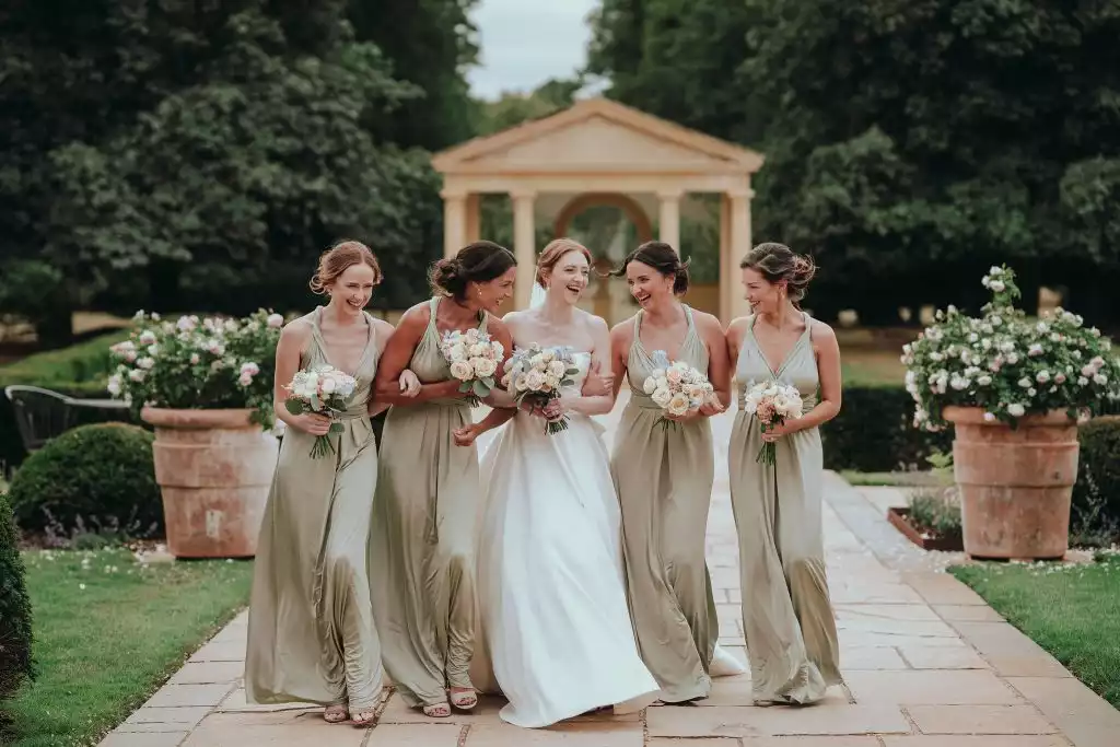 Bride in a strapless gown walking arm in arm with bridesmaids in sage green dresses in front of a garden pavilion ceremony space.