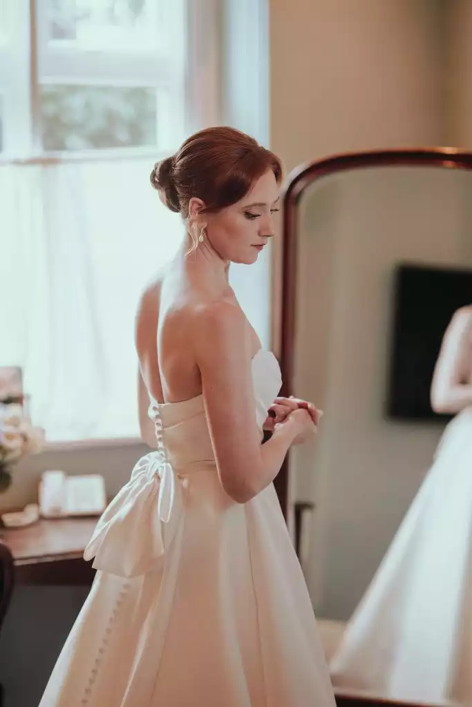 Bride in a strapless wedding dress standing beside a mirror during morning preparations, showcasing the button-back detail of her gown.