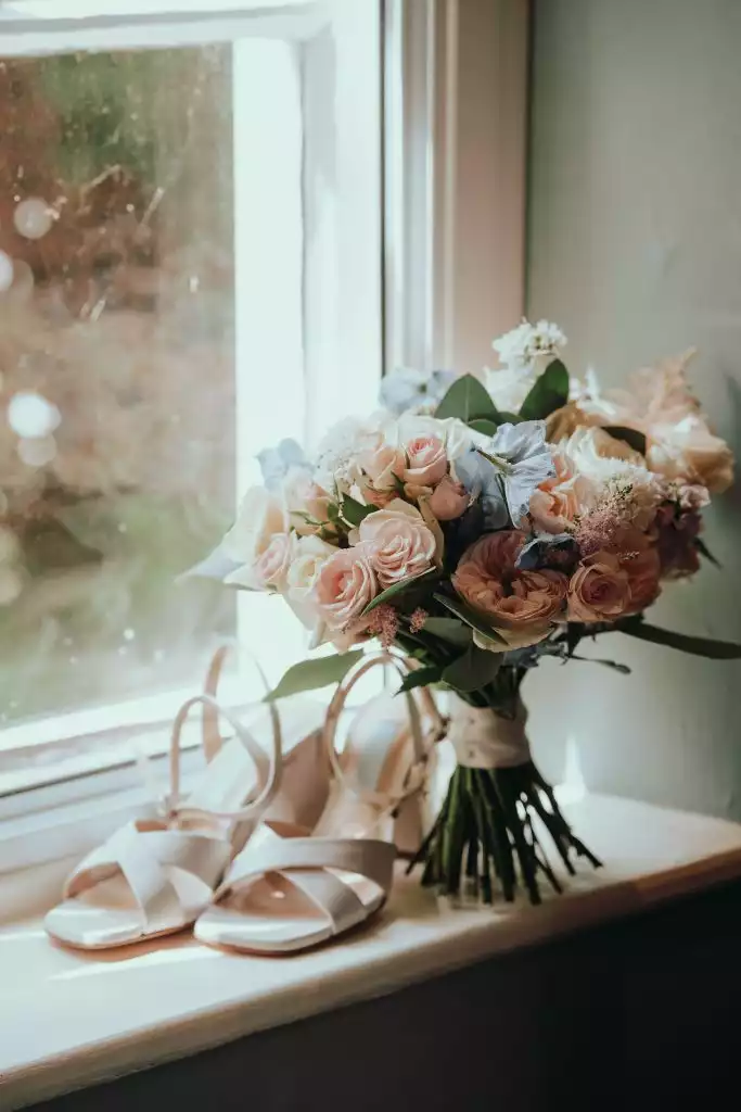 Blush and ivory wedding bouquet resting beside white bridal heels on a windowsill with soft natural light.
