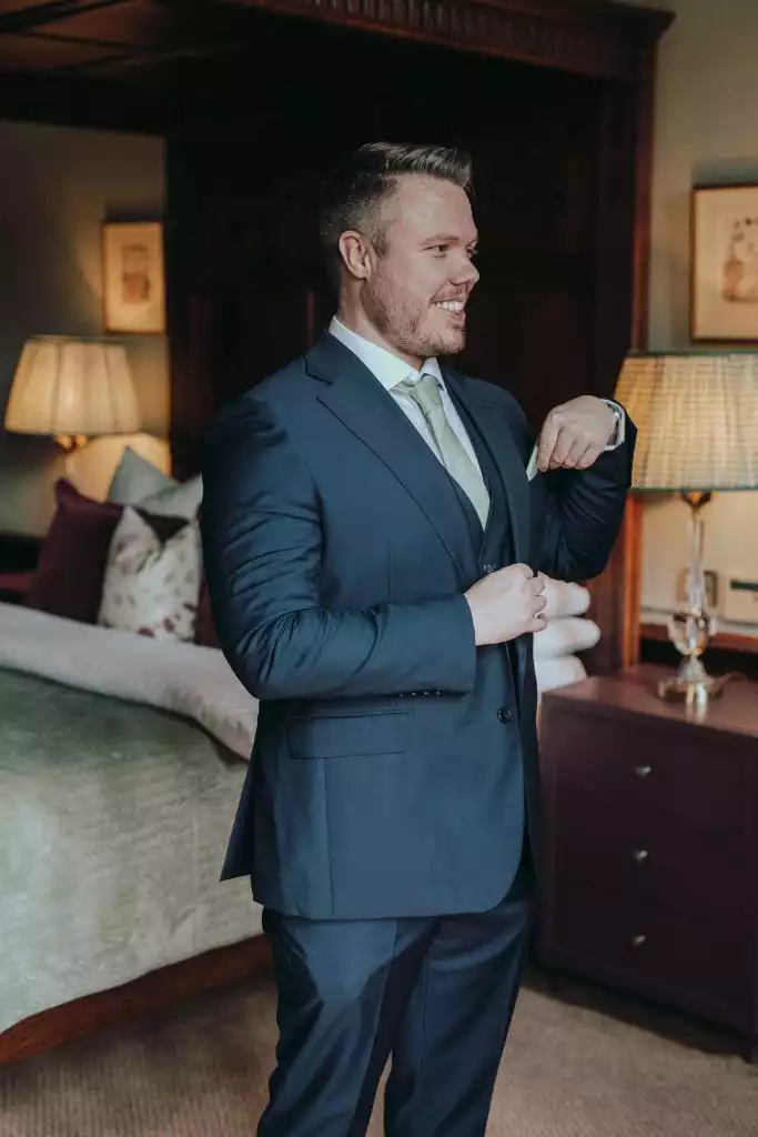 Groom adjusting his tie in a navy suit inside a luxury wedding suite with warm lighting and classic interior décor.