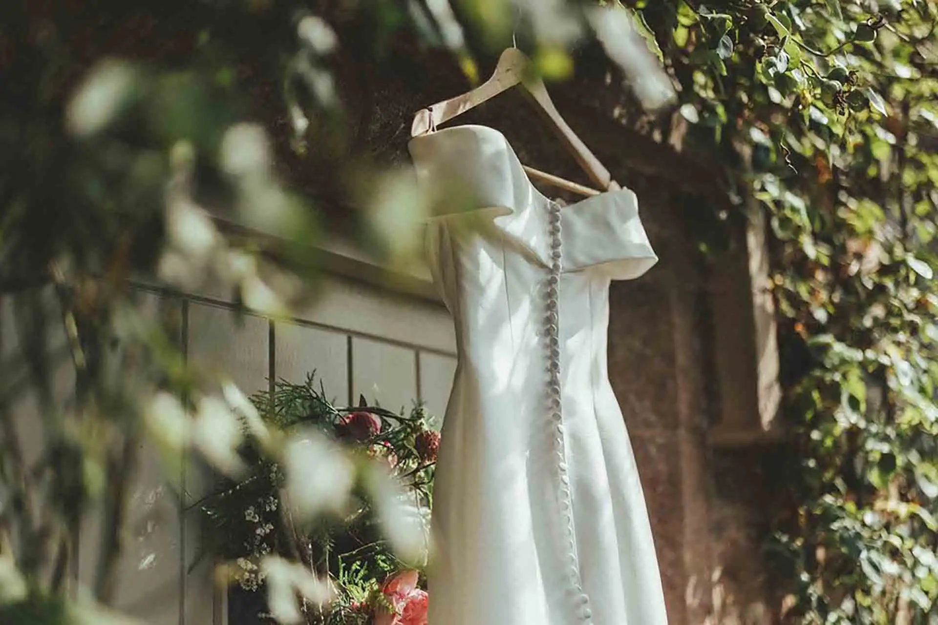 Off-the-shoulder ivory wedding dress hanging outdoors against greenery and stone walls at Rowton Castle.