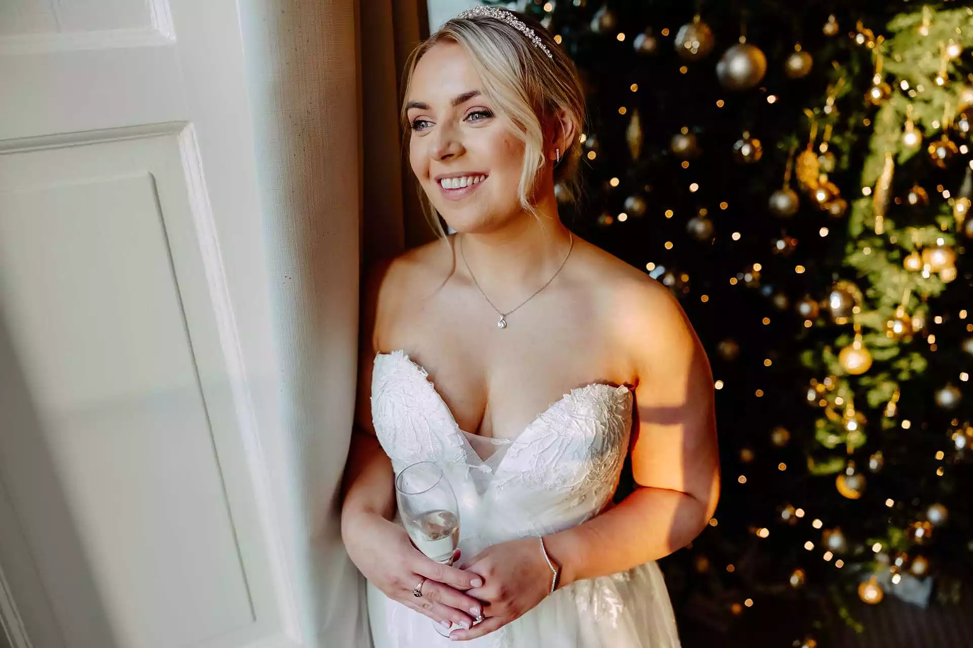 Bride in a strapless wedding gown holding a champagne flute, standing by a softly lit Christmas tree at Rowton Castle.