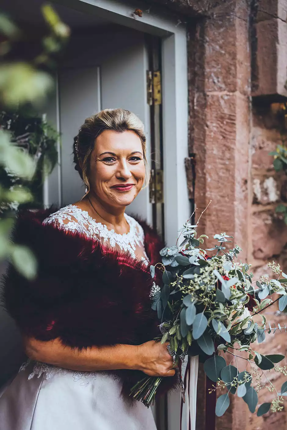 Bride wearing a burgundy faux fur stole and holding a winter bouquet outside the entrance of Rowton Castle.