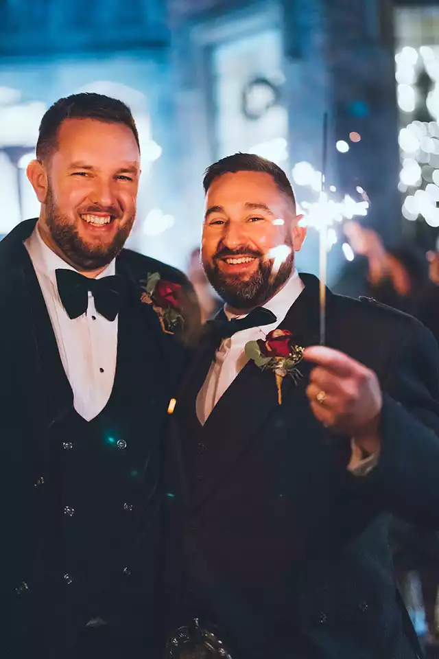 Two grooms in black tie suits smiling and holding sparklers during an evening winter wedding celebration at Rowton Castle.