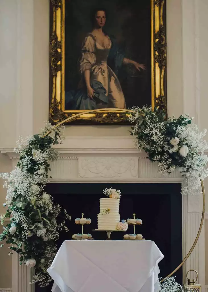Wedding cake display with floral arch and cupcakes inside Rowton Castle