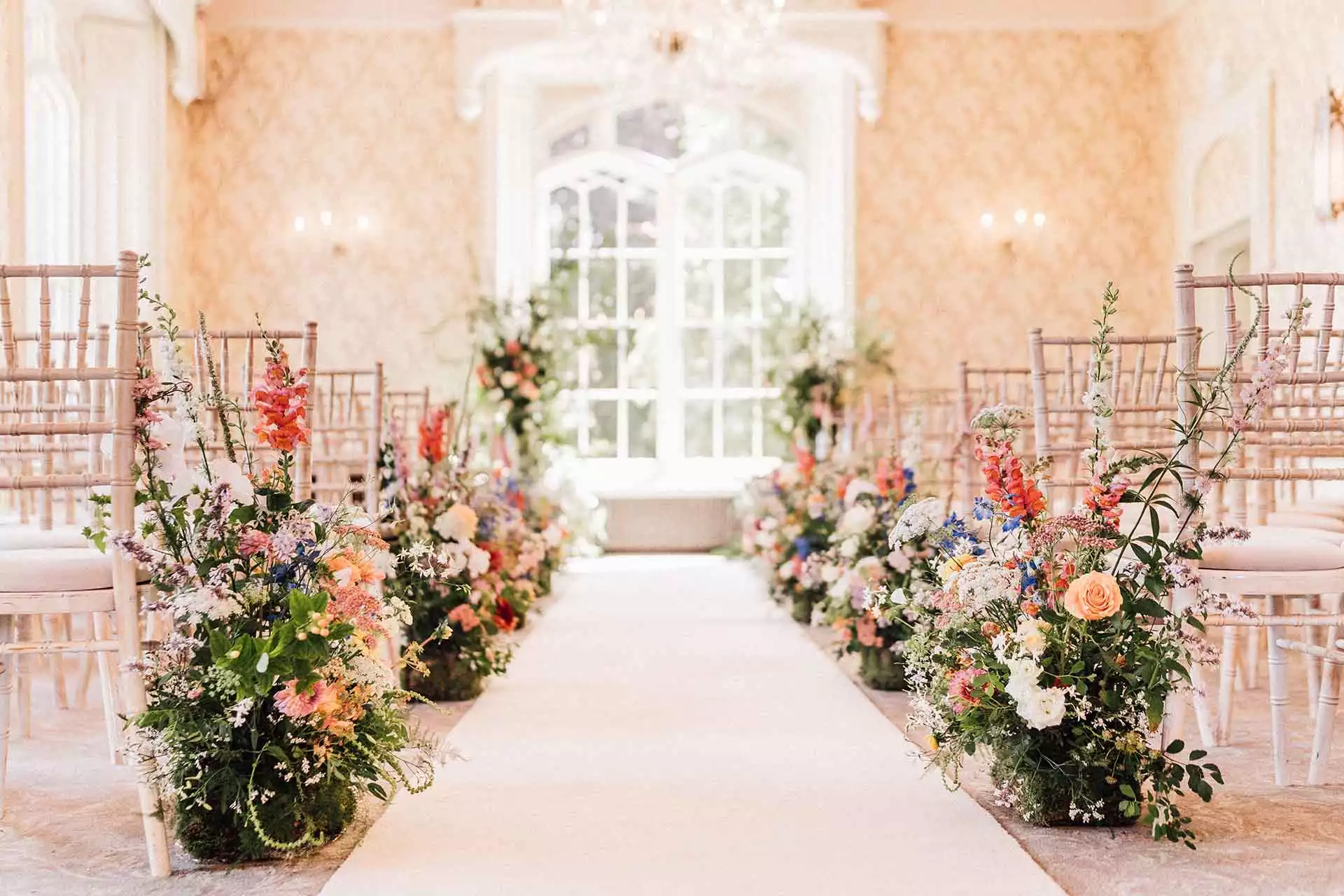 Elegant indoor ceremony aisle lined with colourful floral arrangements and Chiavari chairs at Rowton Castle.
